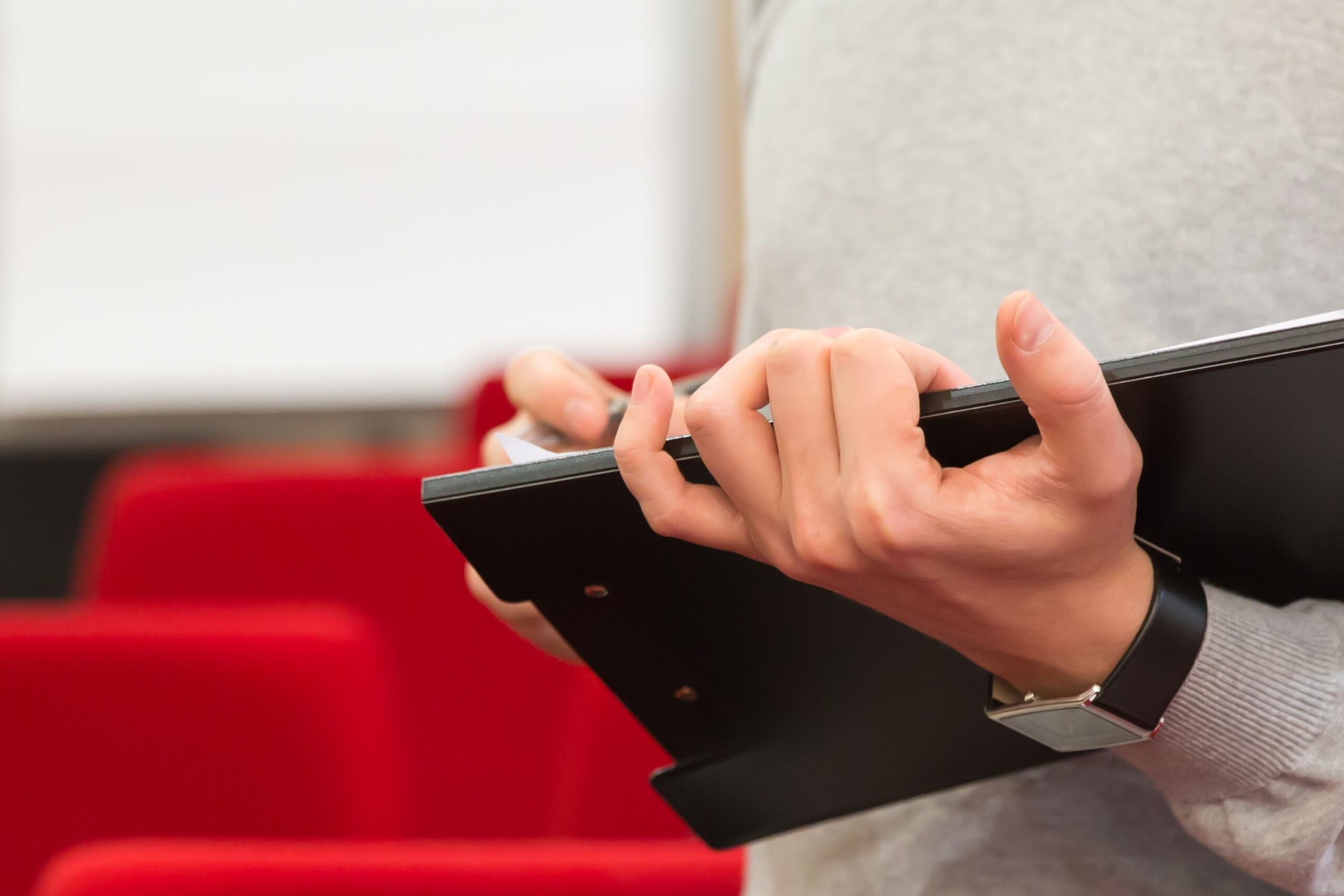Close-up of a person holding a clipboard in a seminar with red seats.